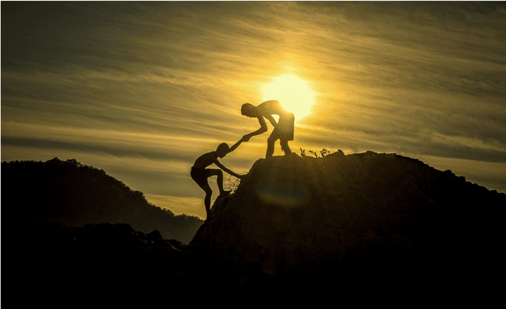 man at the top of a mountaintop reaching hand down to help partner